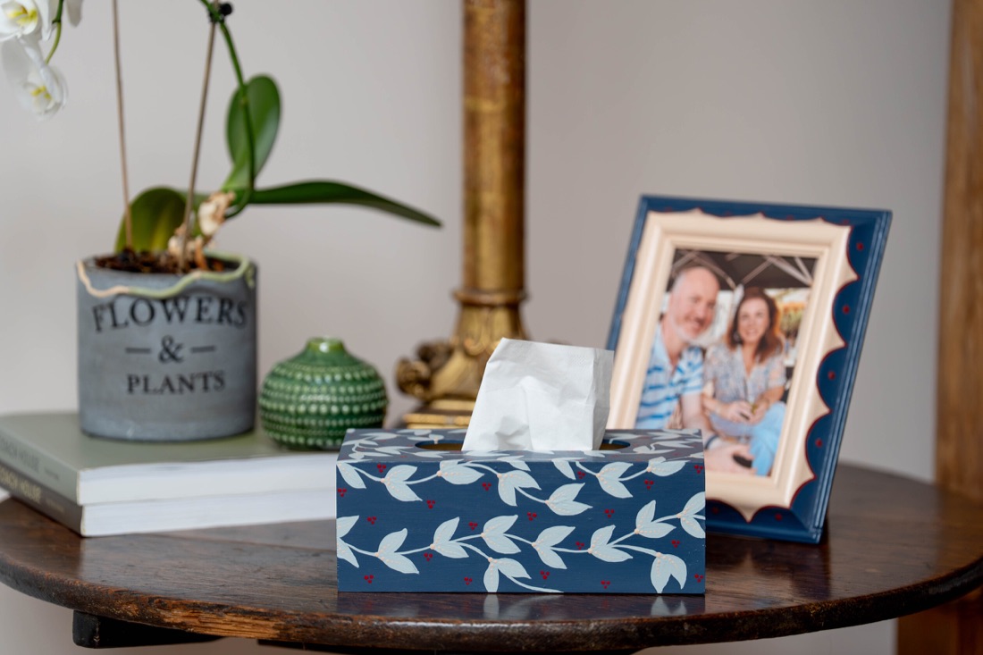 A navy tissue box with hand painted white leaves on a wooden table.